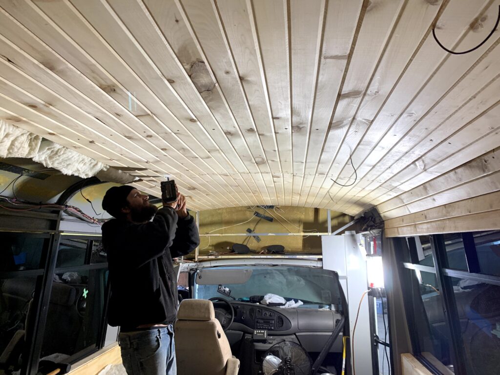 A man holding a power tool, making a hole in the ceiling to install pot lights in our shuttle bus build.