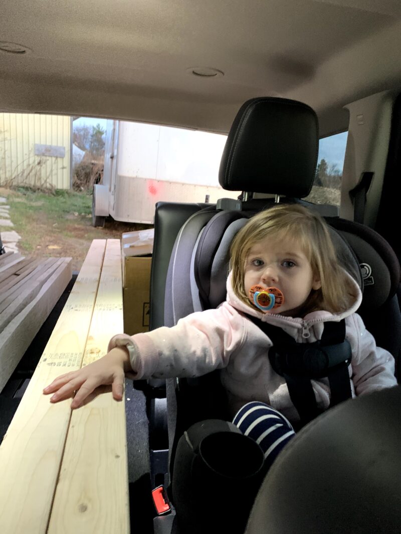 Toddler sitting in her car seat, holding down the lumber we bought to continue our custom bus build.